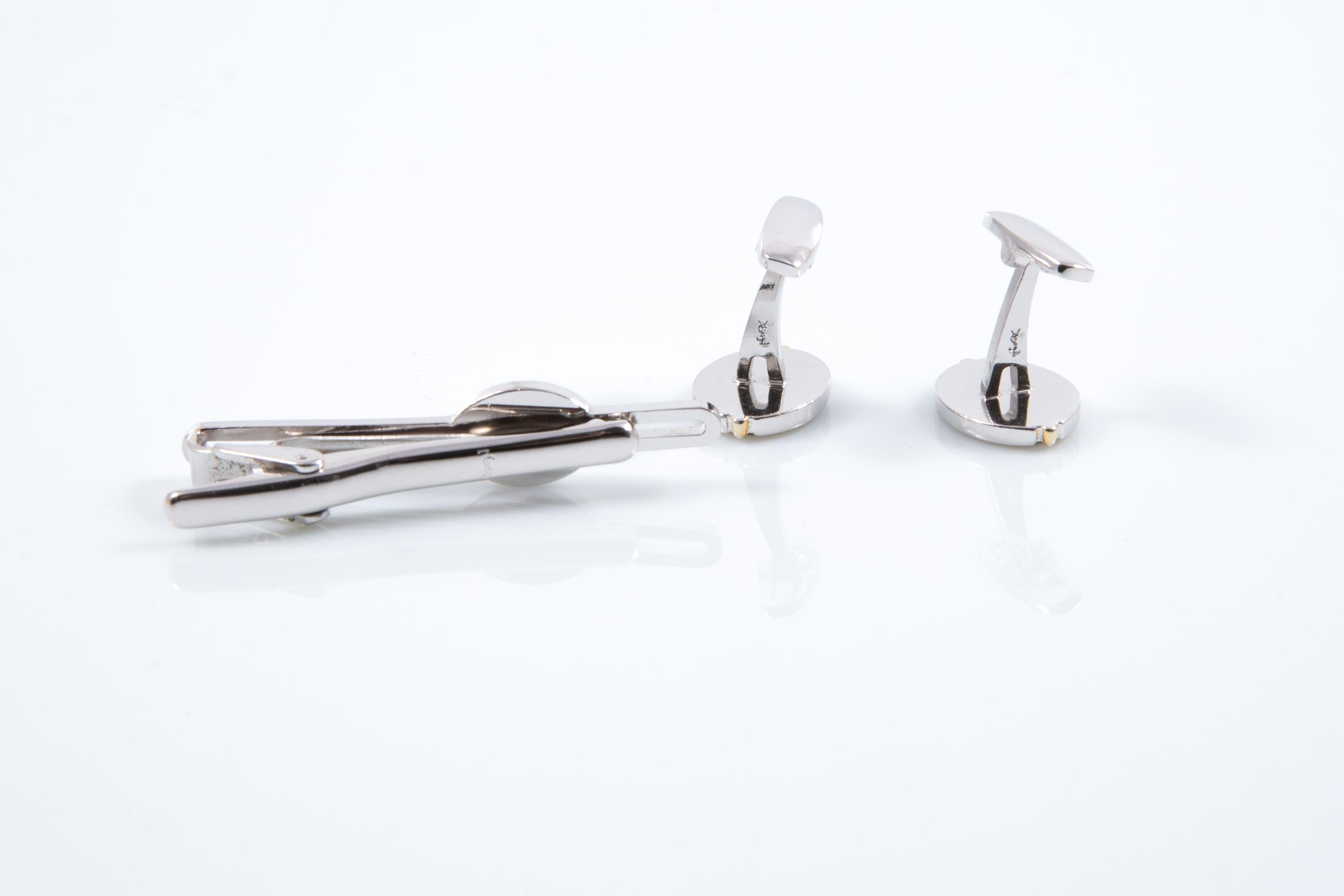 Yves Saint Laurent Silver-tone “YSL” Cassandre cuff-links and tie clip logo link set featuring an oval shape, a center grey stone with a gold-tone YSL & silver-tone outline. Vienen como un par + un accesorio de corbata. 
En buen estado vintage Circa