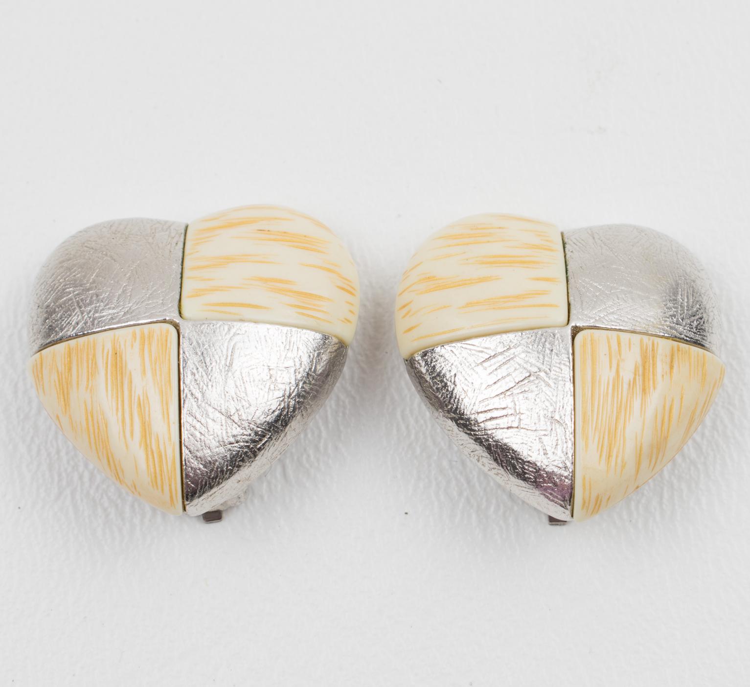 Questi eleganti orecchini a clip firmati Yves Saint Laurent Paris sono caratterizzati da una romantica forma a cuore in metallo argentato con motivo a scacchiera, contrastato da elementi in resina bianca e beige che imitano un motivo a ossa. Ogni
