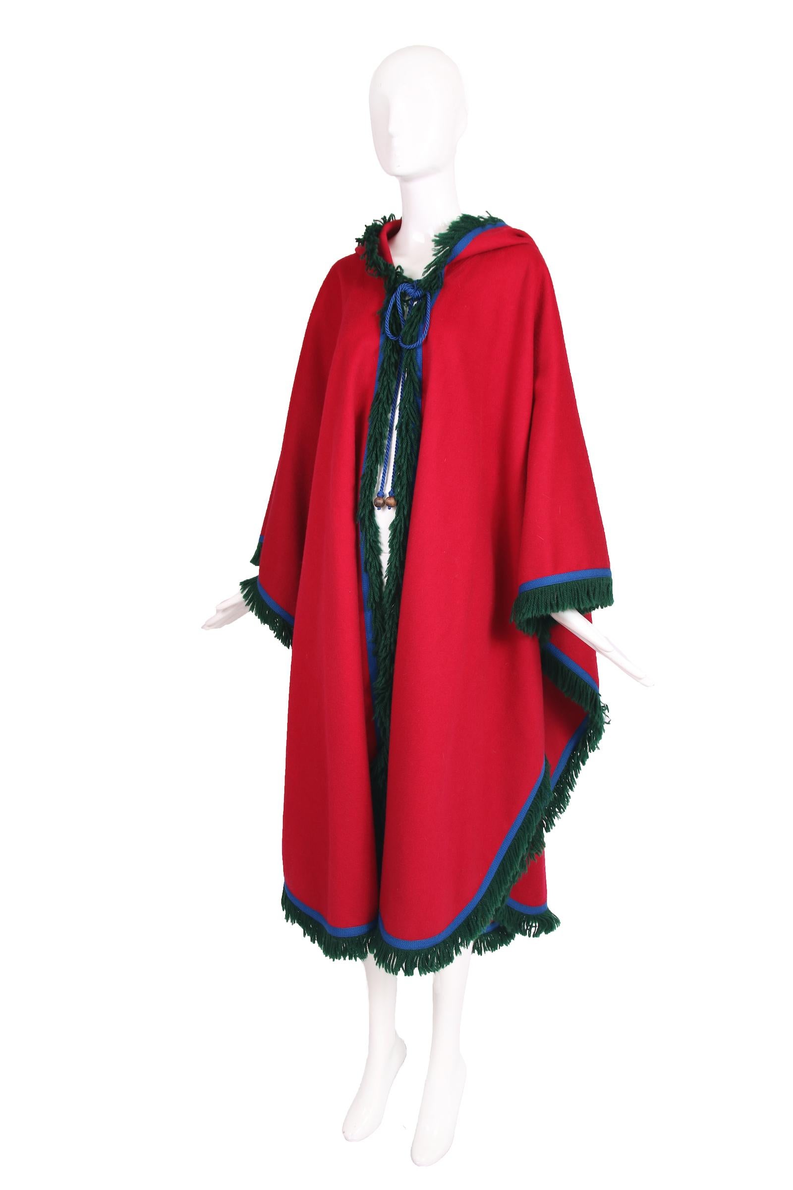1970's Yves Saint Laurent (Rive Gauche) 100% red melton wool cape with royal blue wool cord as trim, green wool fringe and blue silk cord ties at the neck. Cape features a hood with a blue silk tassel attached to the tip. No size tag - one size fits