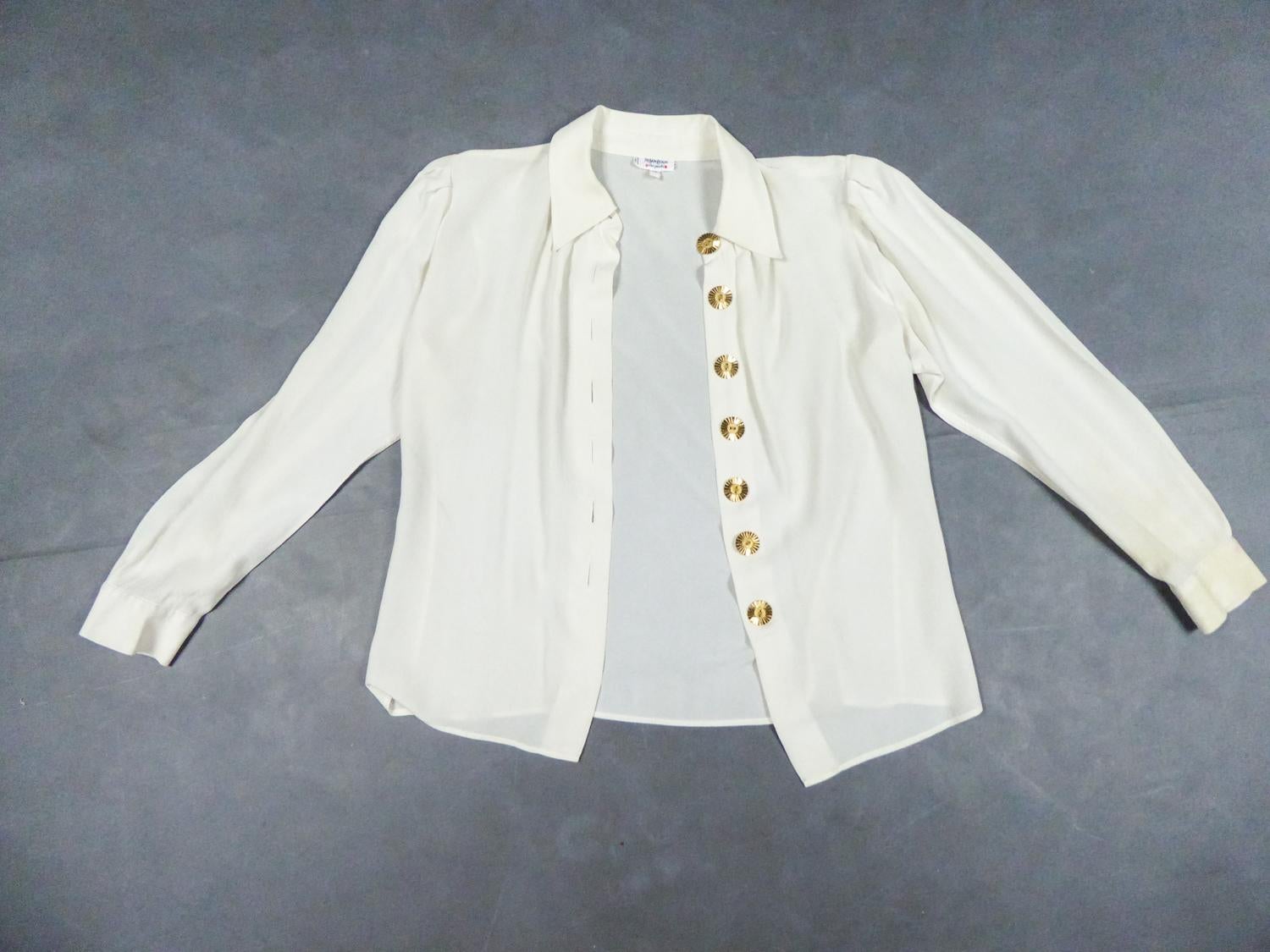 Circa 1990/2000
France

Blouse in cream silk crepe by Yves Saint Laurent Rive Gauche from the years 1990/2000. Classic collar, long sleeves, fluid and loose cut, the blouse closes at the front by buttons in chiseled golden brass recalling suns.