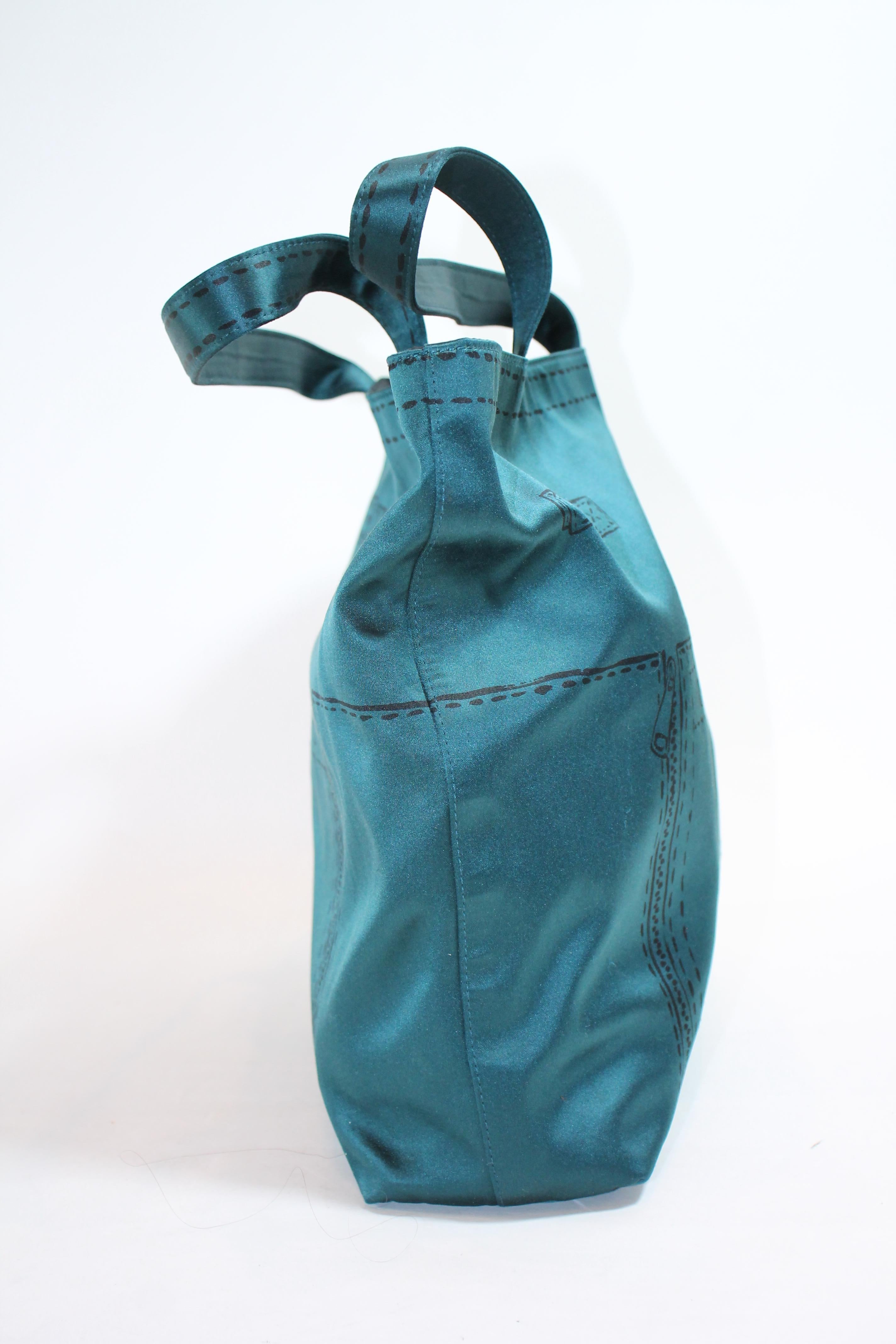 Teal satin featuring the marks of signature side belts and buckles, vertical zippers, and facing pocket. Open top. Dual flat top handles. Satin interior. Two interior opened pockets. One large zippered pocket.