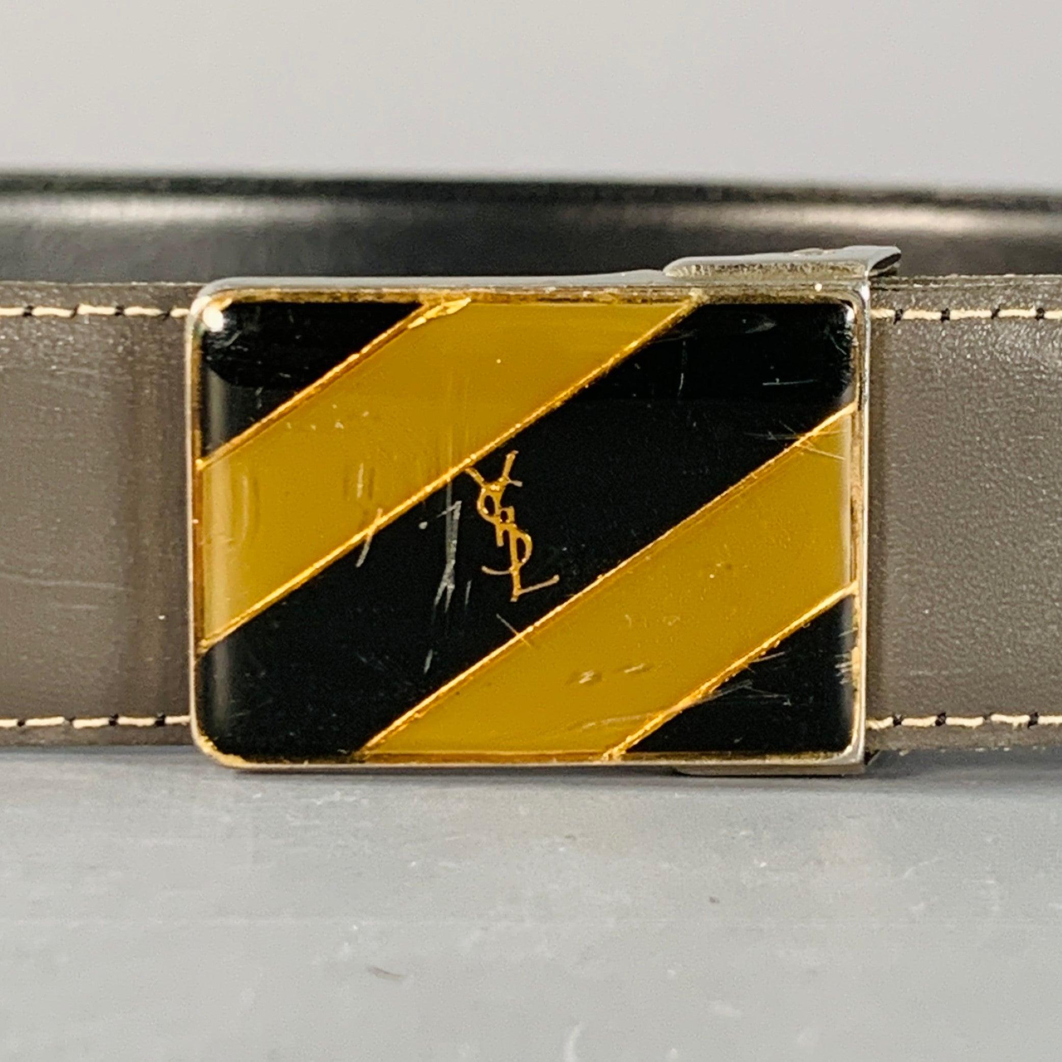 Vintage By YVES SAINT LAURENT
Gürtel aus grauem Leder mit rechteckiger silberfarbener Metallschnalle und goldfarbenem Metall-Logo auf diagonalem Streifenmuster. Hergestellt in Italien, guter gebrauchter Zustand. Gebrauchsspuren an der Schnalle.