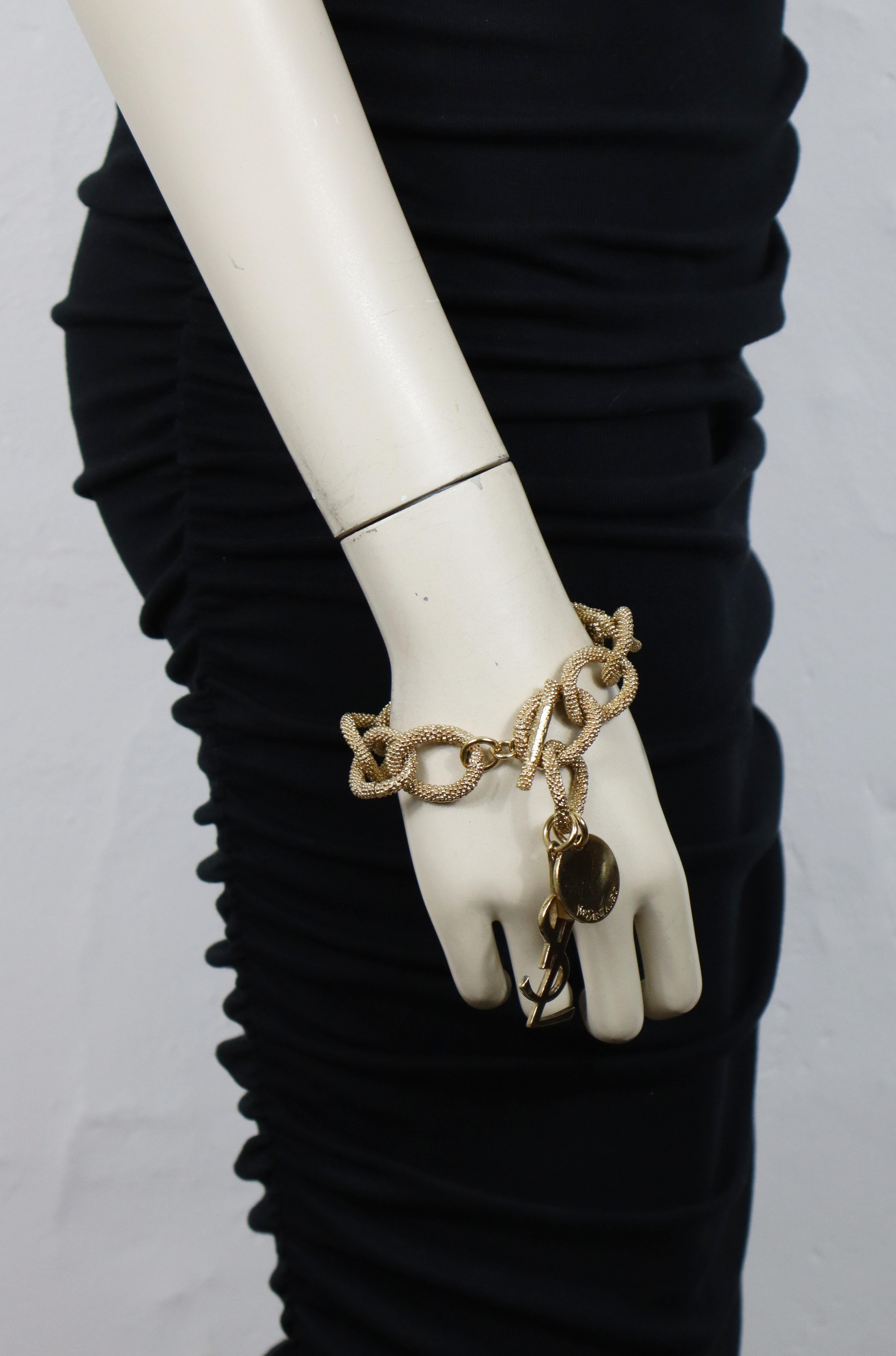 Bracciale a catena YVES SAINT LAURENT in tonalità oro con maglie ovali testurizzate, logo YSL e charm a forma di moneta con impresso YVES SAINT LAURENT.

Chiusura con barra a T.
Lunghezza regolabile.

Yves Saint Laurent in rilievo sulla barra a T e