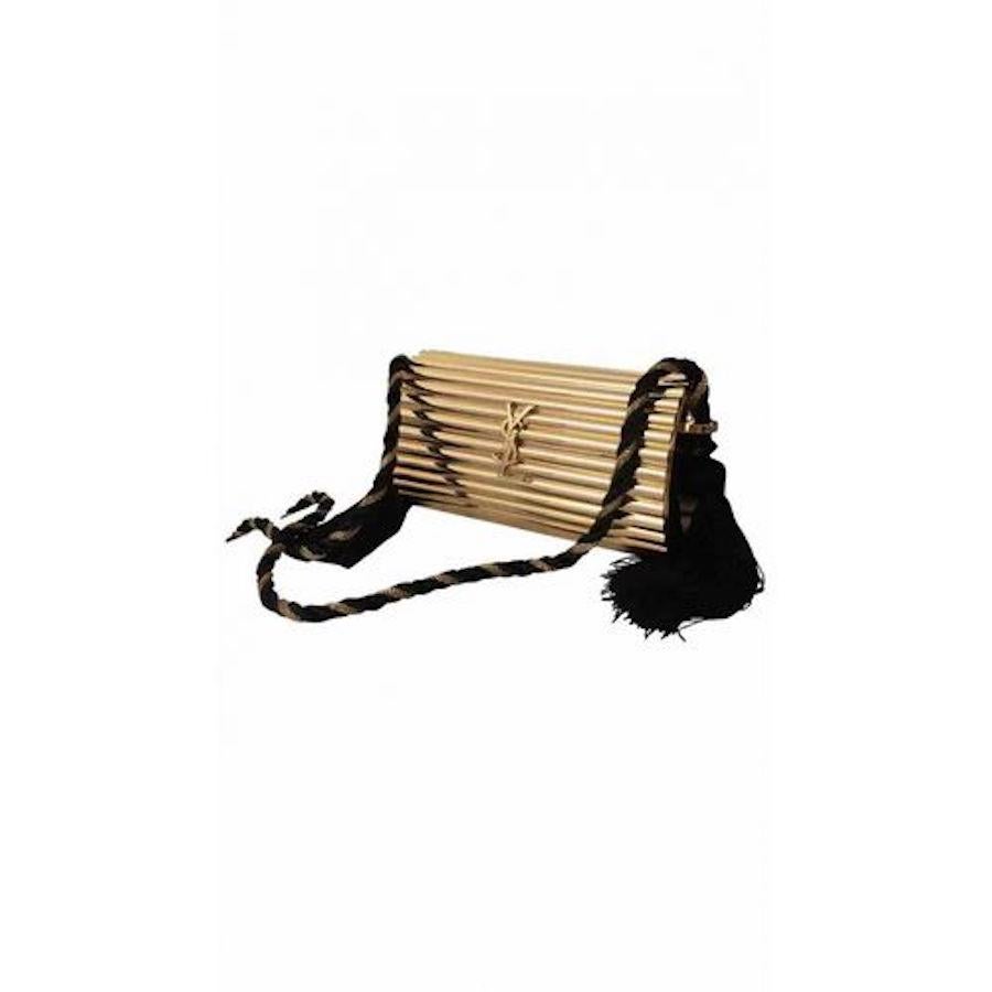 This iconic YSL bag from the 1980’s has a rigid cylindric exterior made from shiny ribbed gold-tone metal. The bag features a long black and gold braided silk cord shoulder strap, dual black silk tassels on both ends of the bag, an interlocking YSL