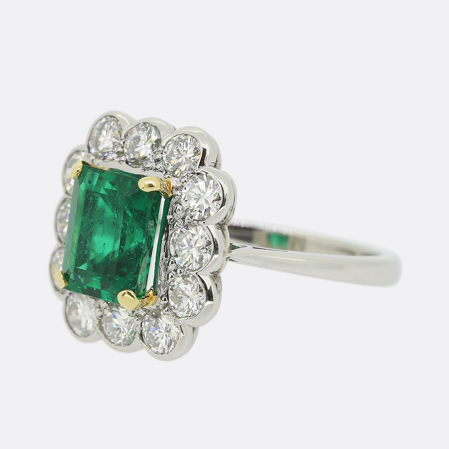 This is a wonderful emerald and diamond cluster ring. The ring features a central emerald cut Zambian emerald which possesses a beautifully intense green tone which is highly complimented by the bright white round brilliant cut diamonds which
