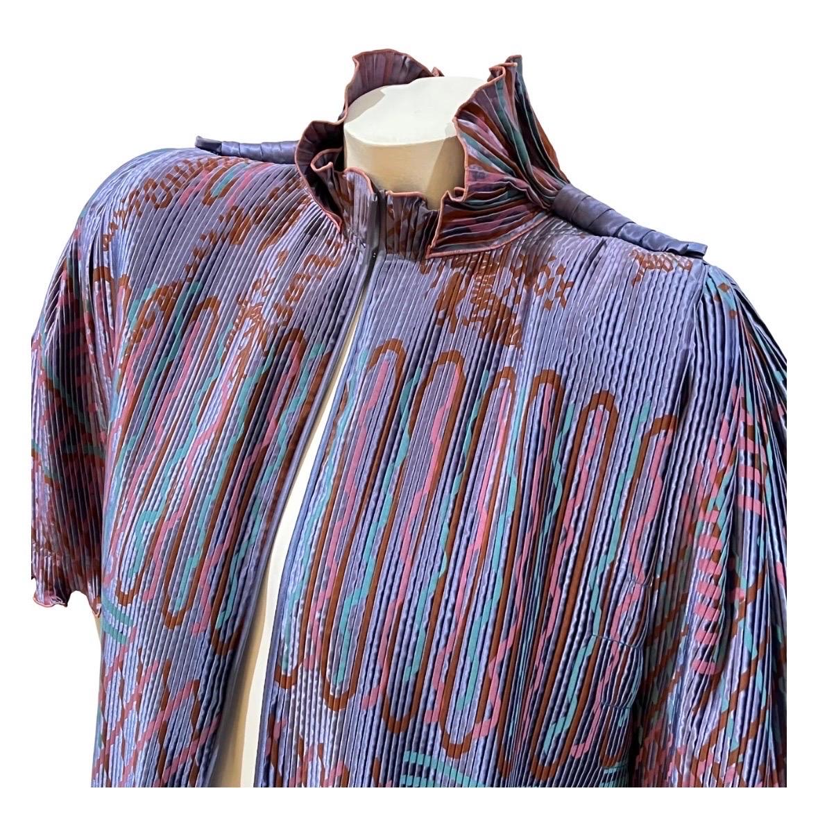 Pleated capelet top by Zandra Rhodes
Circa 1980's
Made in England
Periwinkle w/ turquoise, pink and orange line designs throughout 
Pleated fabric 
Ruffled collar and hems
Single hook closure at neck 
Short, fan sleeves
Shoulder pads 
Side slits
