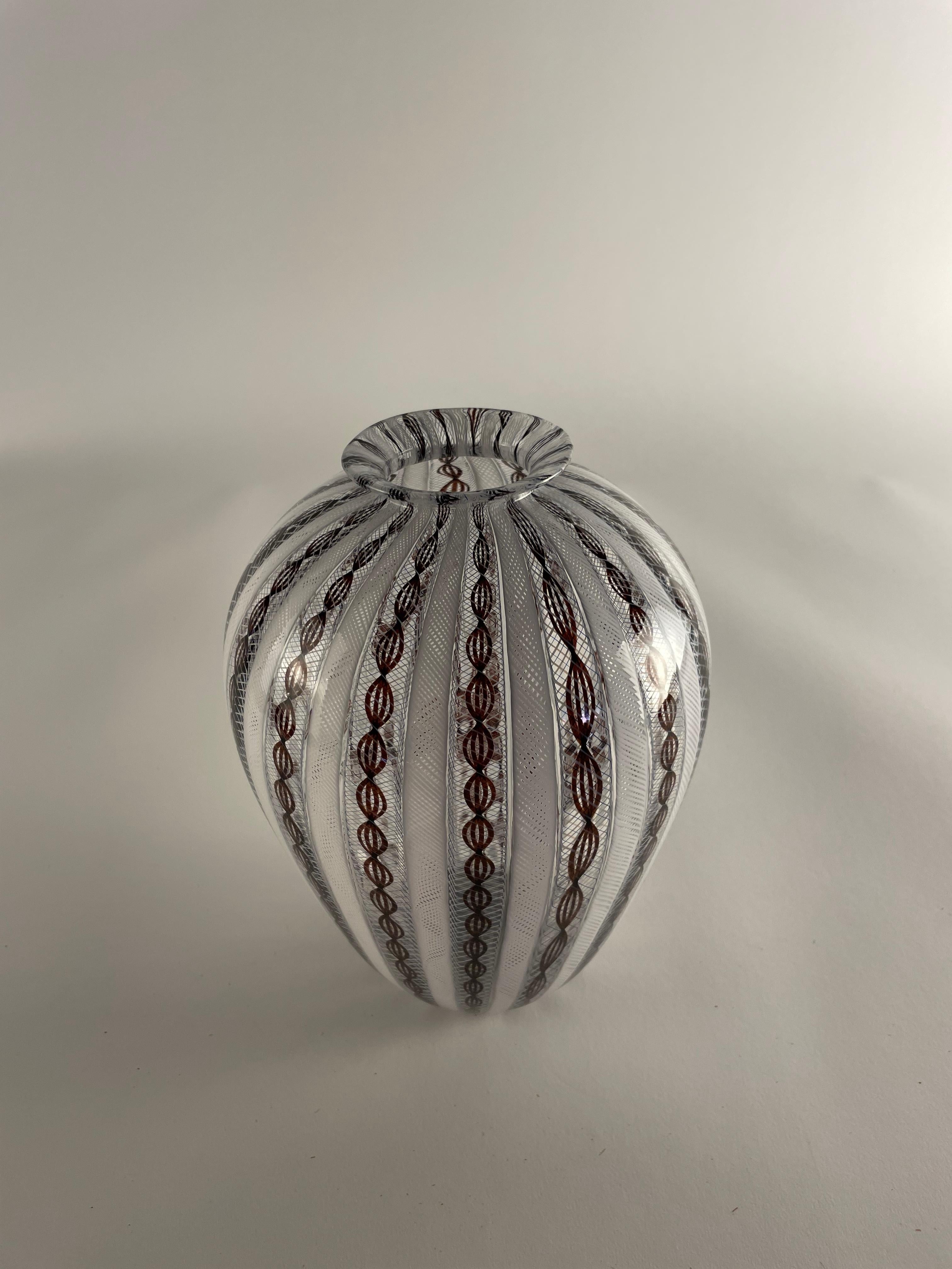 Voici ZANFIRICO, un chef-d'œuvre de l'artisanat du verre de Murano. Ce vase exquis présente l'art du canne zanfirico, une technique complexe qui entrelace plusieurs fils de verre pour créer un motif hypnotique semblable à de la dentelle. Seuls les