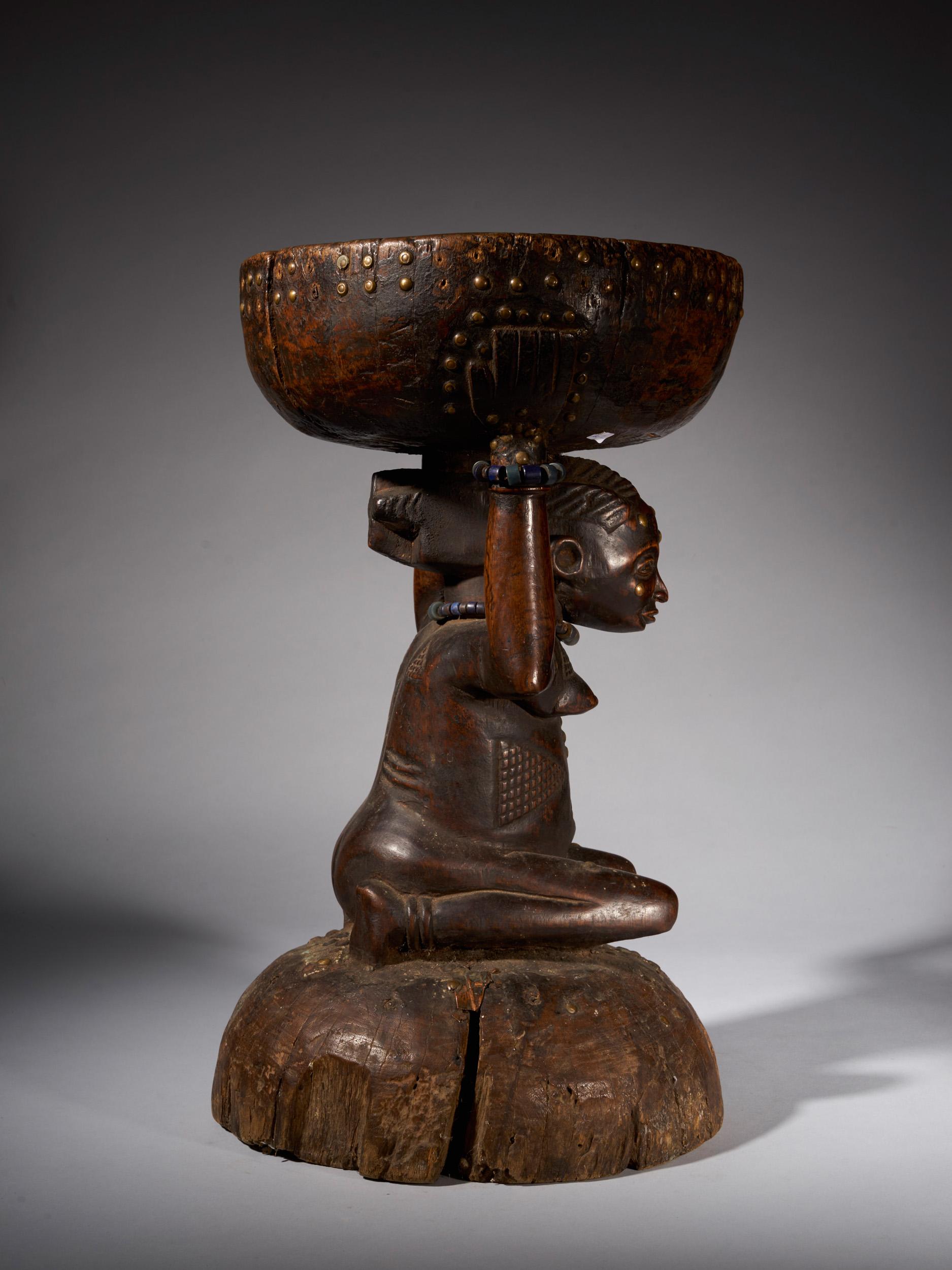 The stool represented here is a typical Caryatide stool of the Zela people in the Democratic Republic of Congo. The stool is carved out of wood and is held up by a kneeling woman covered with typical Zela scarifications, showing her high ranking