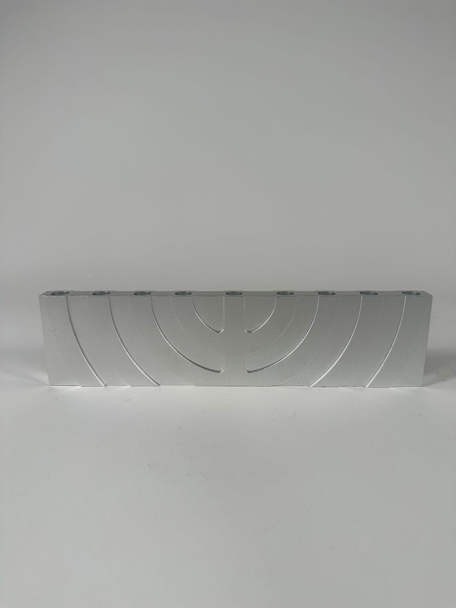 This striking Hanukkah lamp, crafted by the renowned Israeli artist Zelig Segal, exemplifies a fusion of tradition and modernist design. Made from aluminum, the lamp features a minimalist, geometric interpretation of the classic menorah, with a