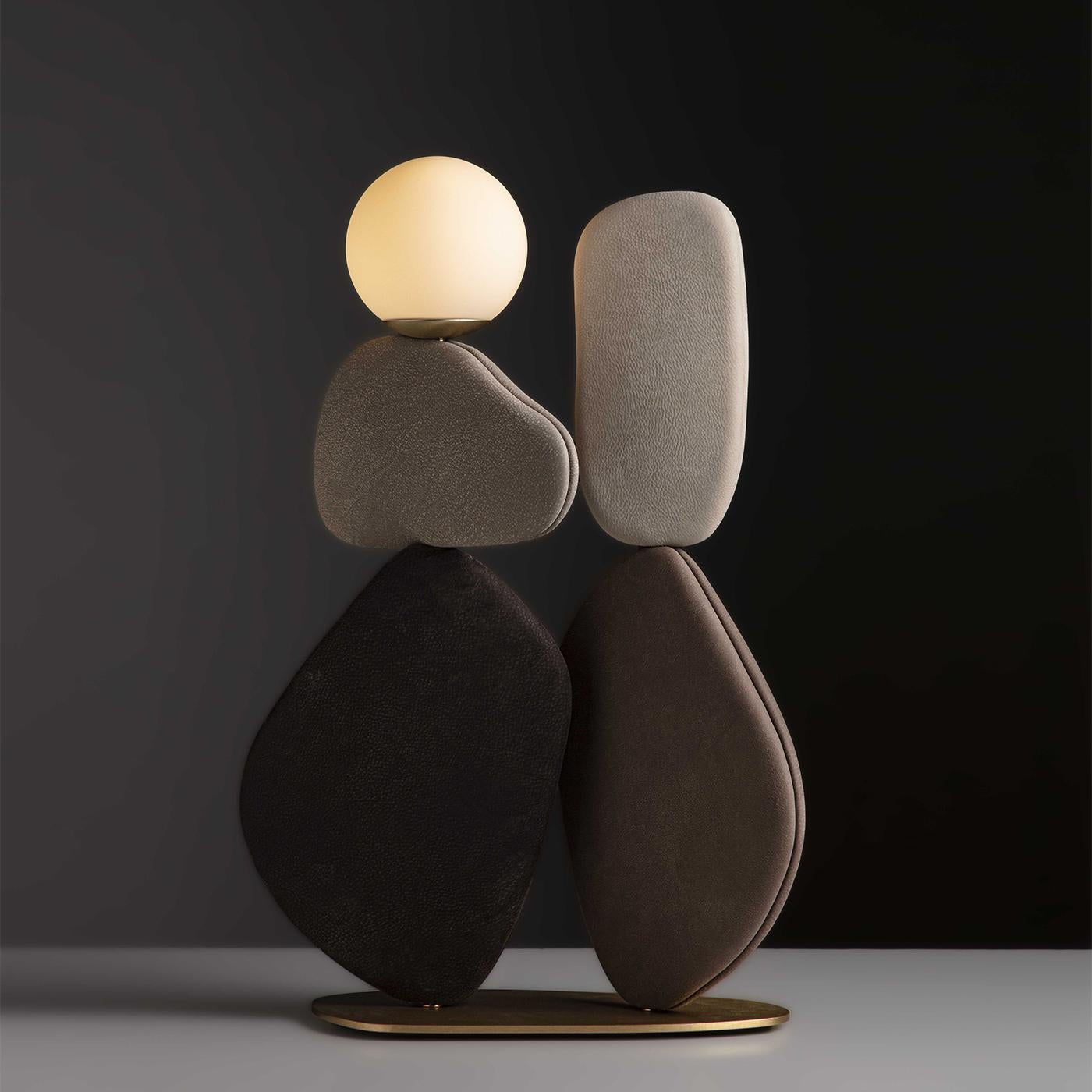 Una lampada da terra scultorea caratterizzata da una base in ottone che sostiene volumi dolcemente curvi in pelle marrone, ispirati all'arte del bilanciamento della pietra. Questi elementi a forma di petalo possono essere ruotati per creare varie