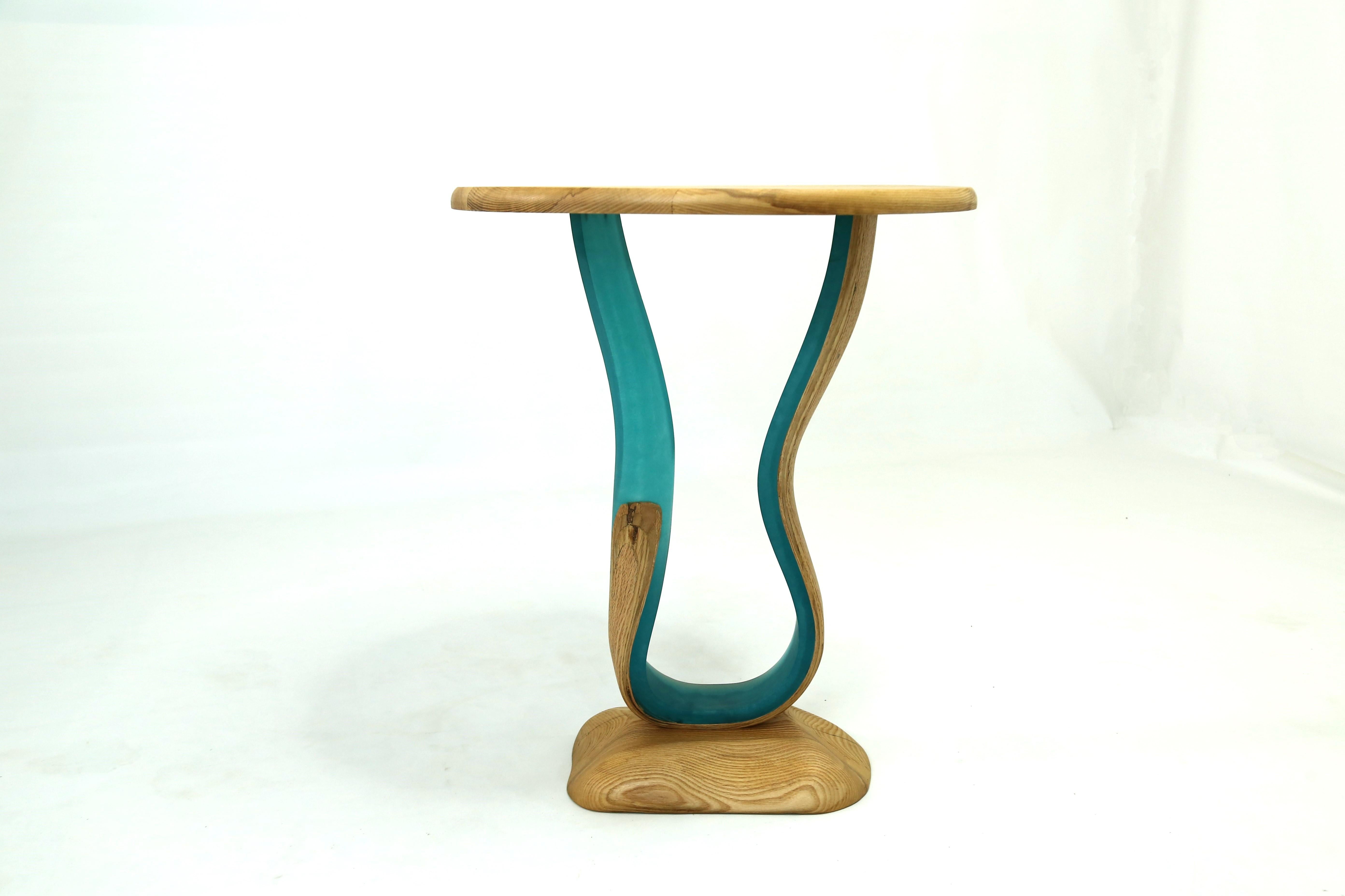 La table Zeta est une table d'appoint dotée d'un plateau circulaire en frêne et d'une base sculptée de forme libre. Le corps de la pièce est constitué de résine fusionnée avec des bandes de bois de frêne qui, ensemble, se tordent et tournent,