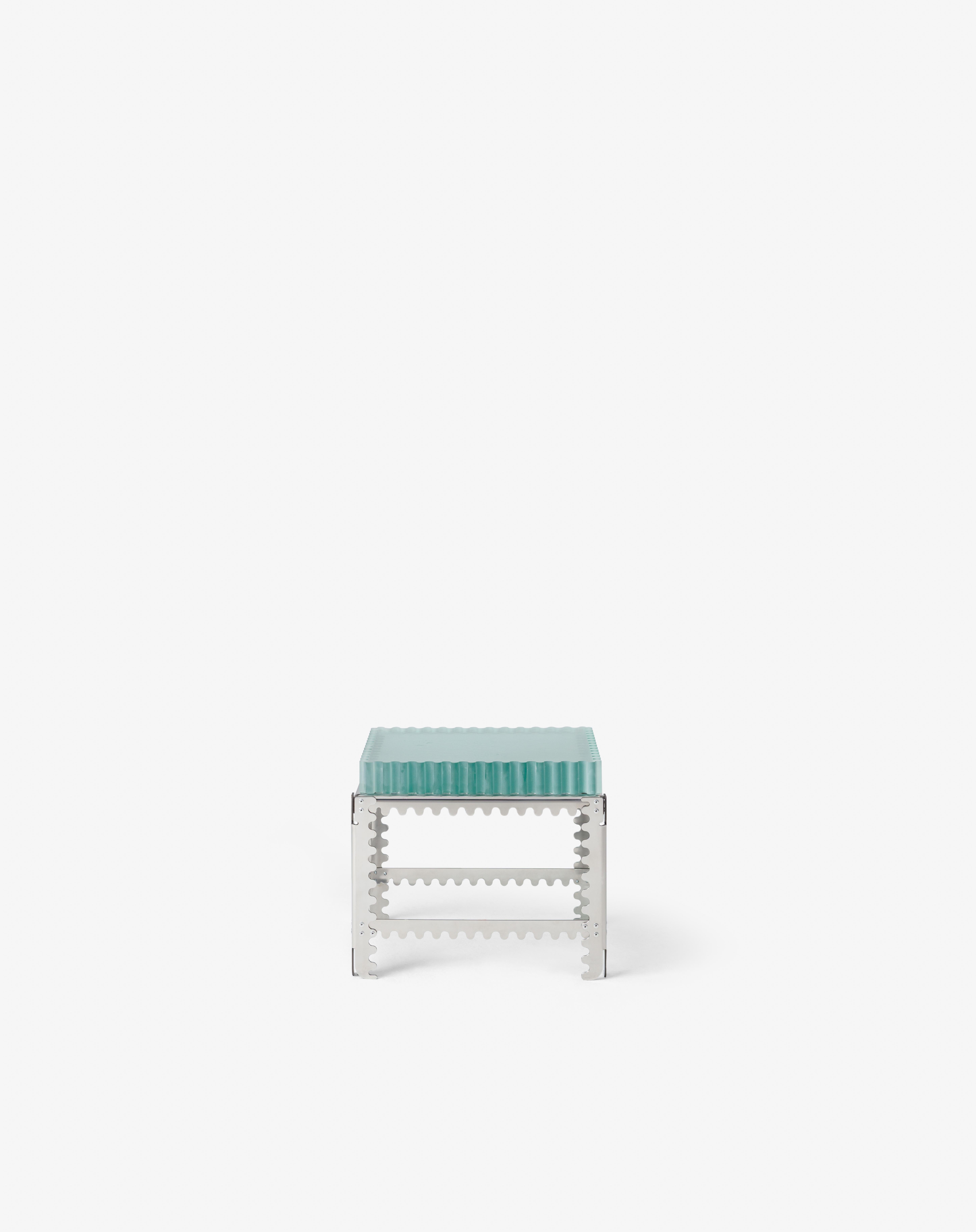This low table pairs cast glass with a polished stainless steel frame,  resembling a glass plinth suspended in a geometric scaffold. The textured transparency softens the strict grid of the structure, balancing mechanical precision with visual