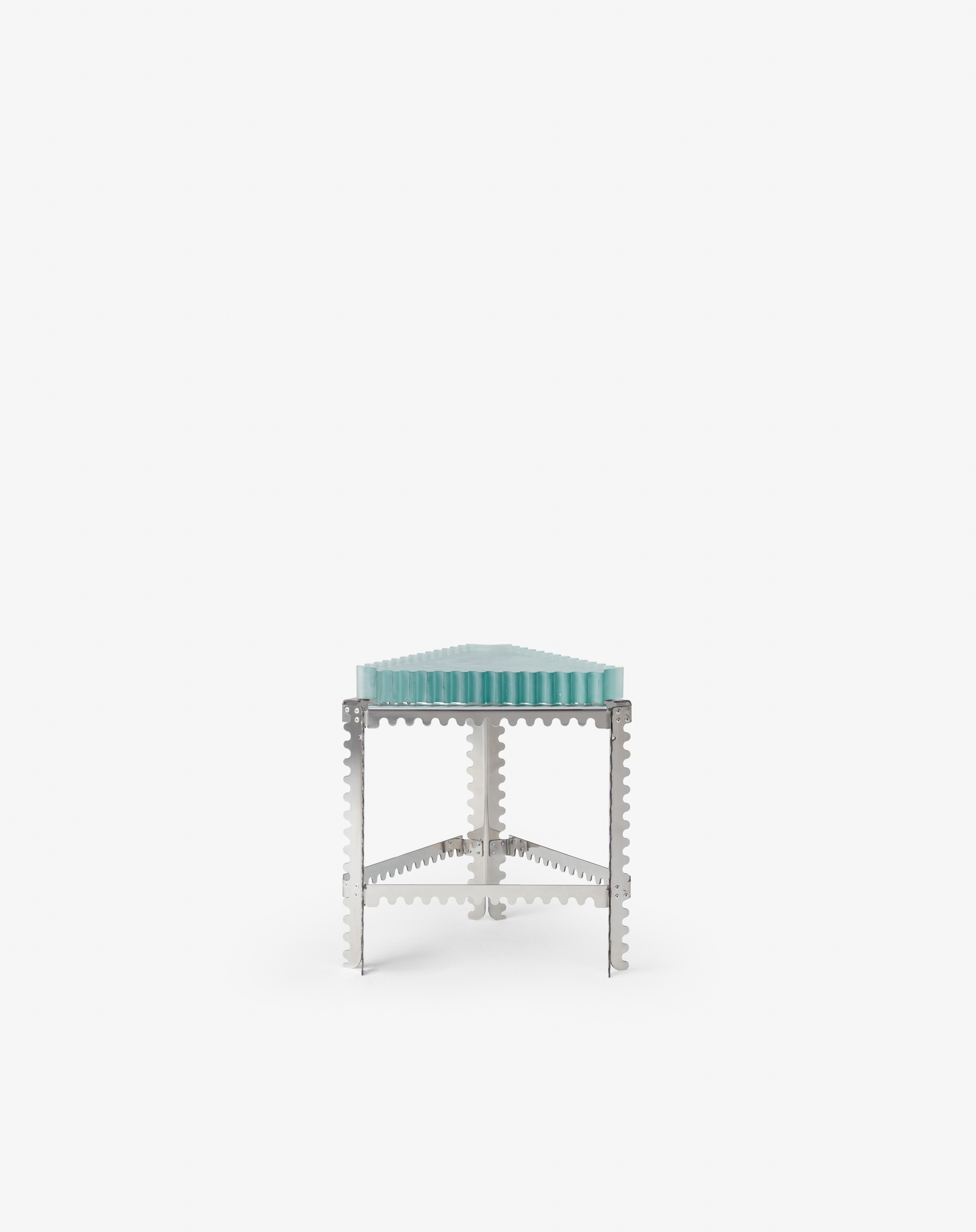 This compact triangular table features a ribbed cast-glass top on a polished stainless steel frame. The jagged metal legs, reminiscent of saw blades or structural bracing,introduce an engineered rhythm. Light refracts through the
glass with shifting