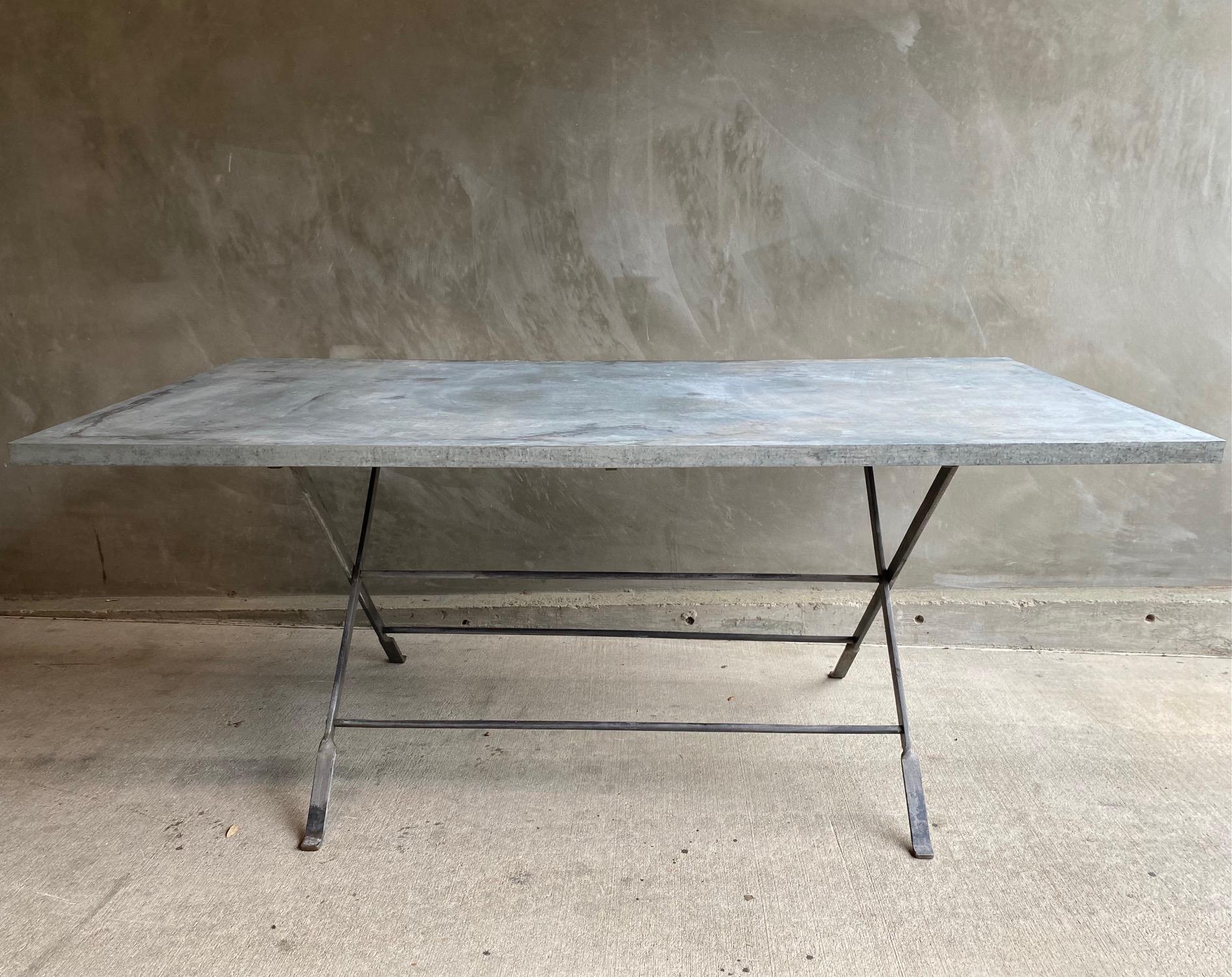 Zinc wrapped dining table top on iron X shape trestle base. Zinc top shows natural patina and variation to the surface. Heavy weight table is very sturdy and appropriate for a primary dining table. Convenient folding mechanism allows for easy
