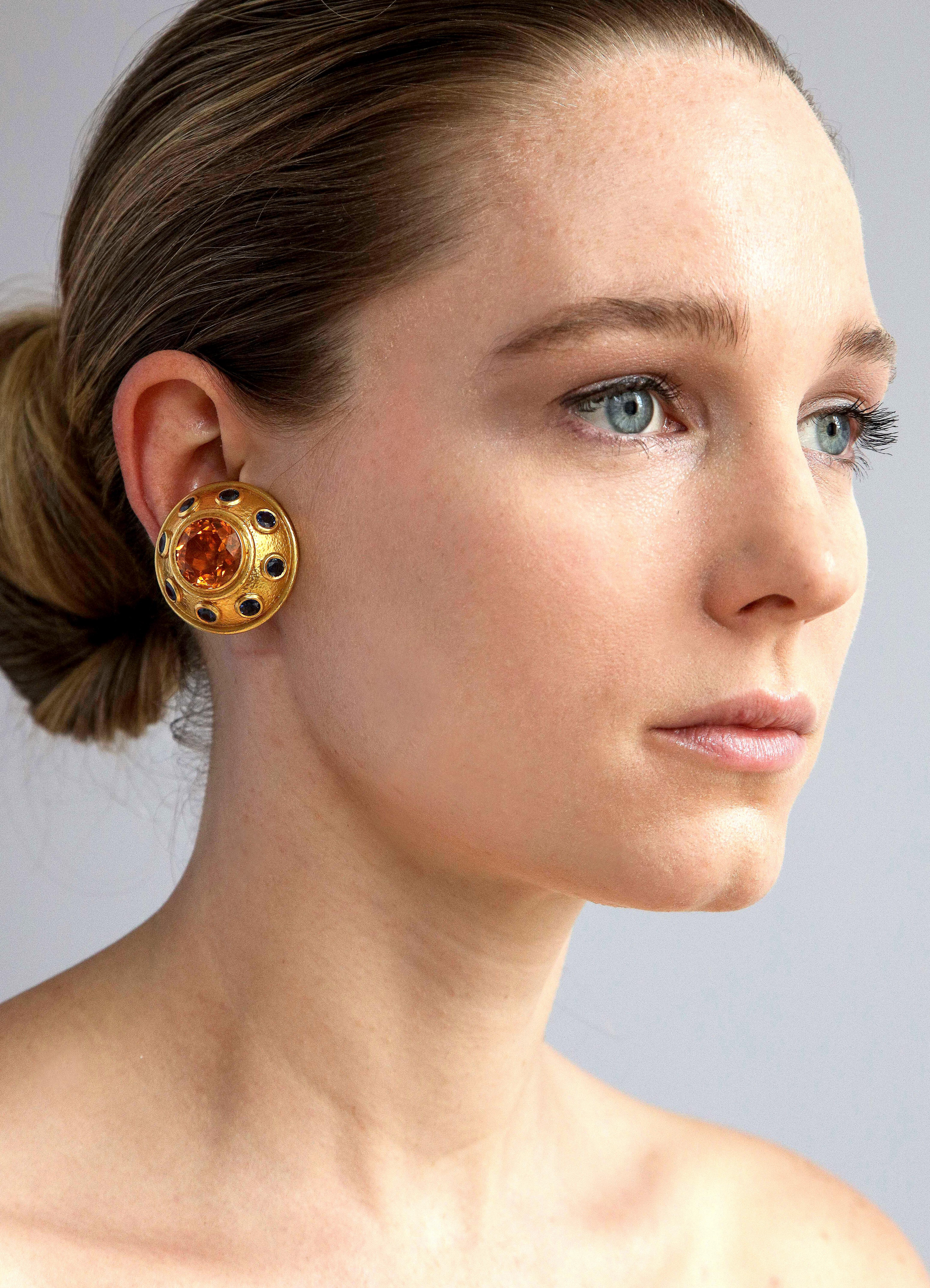 We offer a dramatic pair of Zolotas 22K gold ear clips in a hammered finish set with an impressive golden citrine and ringed in large sapphires. Marked Zolotas and 22K gold. Measuring 1 1/4 inch diameter. Weighing 45.3 grams.