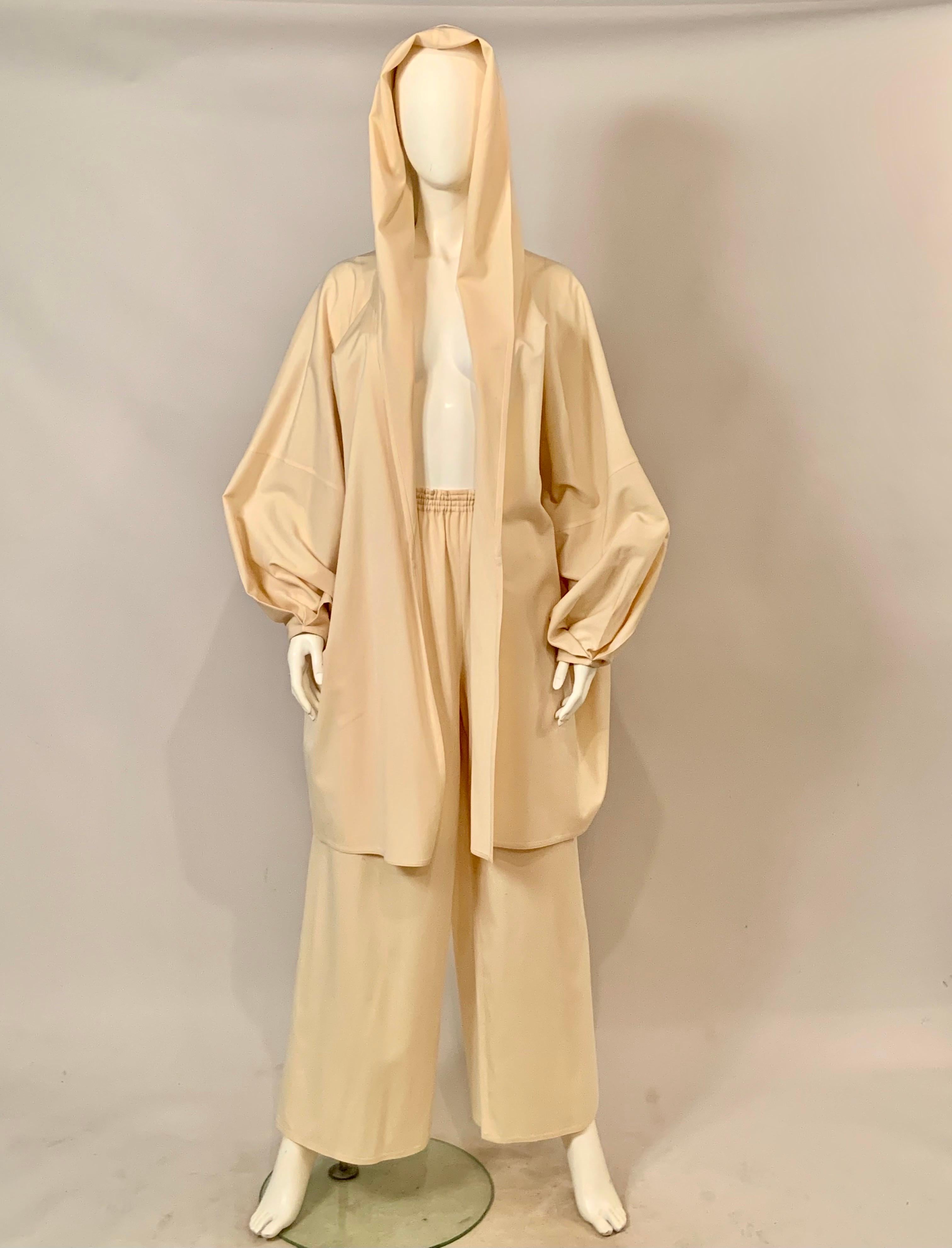  Questo abito in lana color crema di Zoran ha una giacca lunga e ampia con cappuccio e maniche piene con pieghe sui polsini. Il cappuccio della giacca continua lungo la parte anteriore della giacca e forma un bavero. I pantaloni hanno una vita