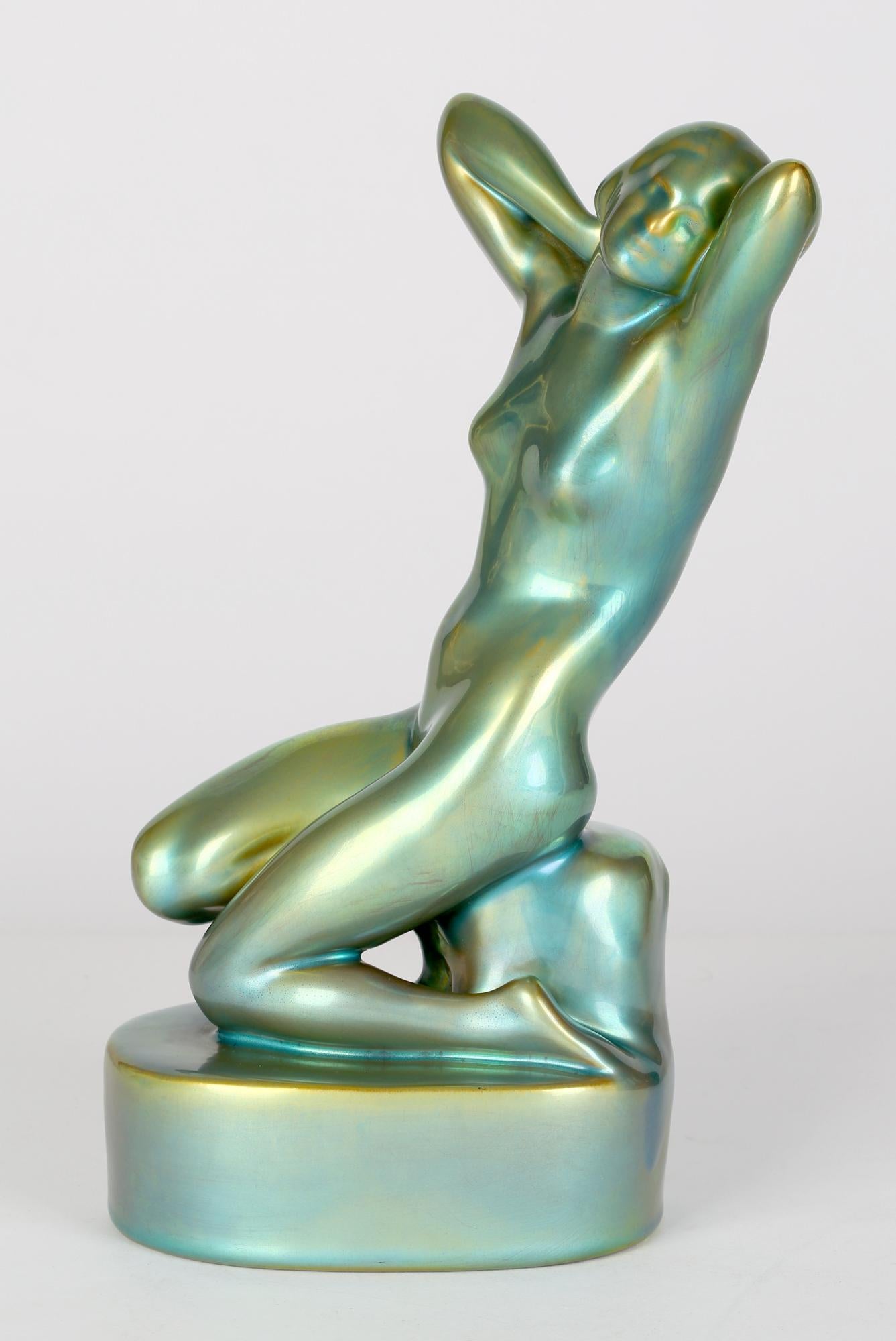 A very fine Art Deco Hungarian art pottery sculptural nude figurine in eosin metallic green glazes dating from around 1930. The figure is mounted on an oval shape base and sits on a rock and kneels on one leg holding her hands behind her head. The