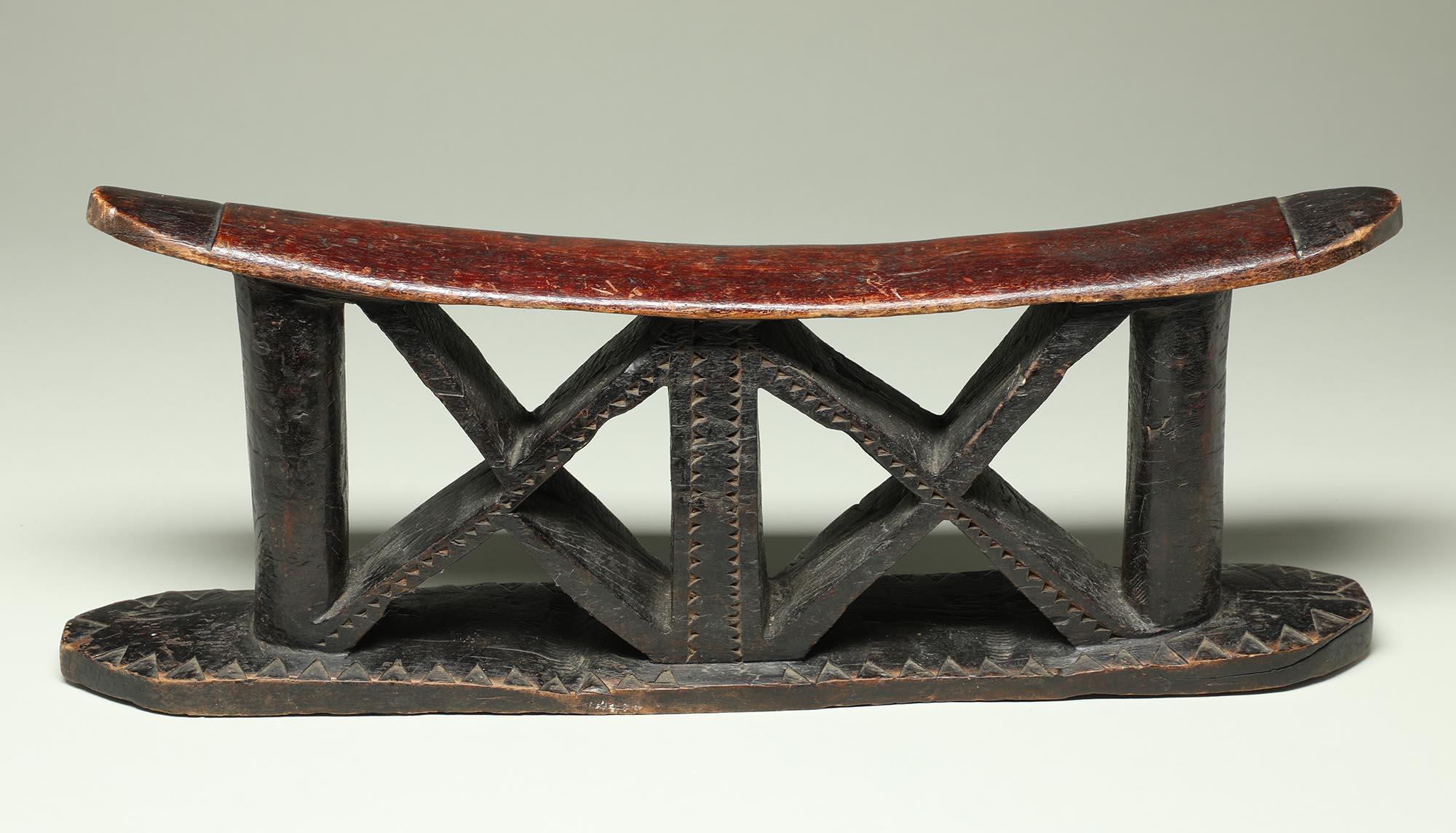 Zulu carved wood double headrest with stylized human figure in middle, from South Africa.
In stylized human form within a pair of vertical columns, with triangle arms and legs and head merging with top.  Red pigment on top.
It is in good condition