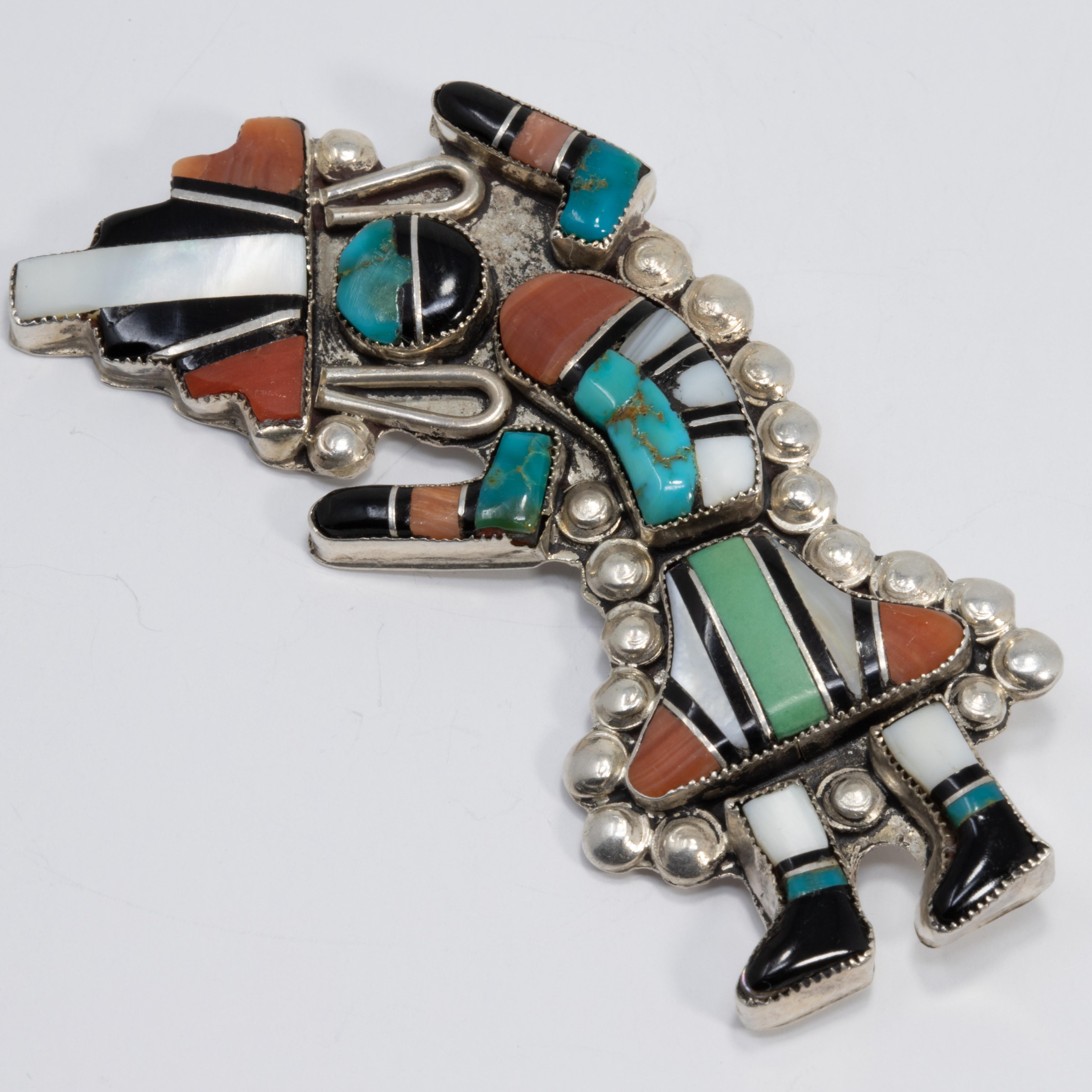 A Native American Zuni dancing man pin featuring mosaic-style turquoise, coral, jet, and mother of pearl inlays bezel-set in a sterling silver setting. A depiction of the symbolic and well-known Indian rainbow god design popular with Native American