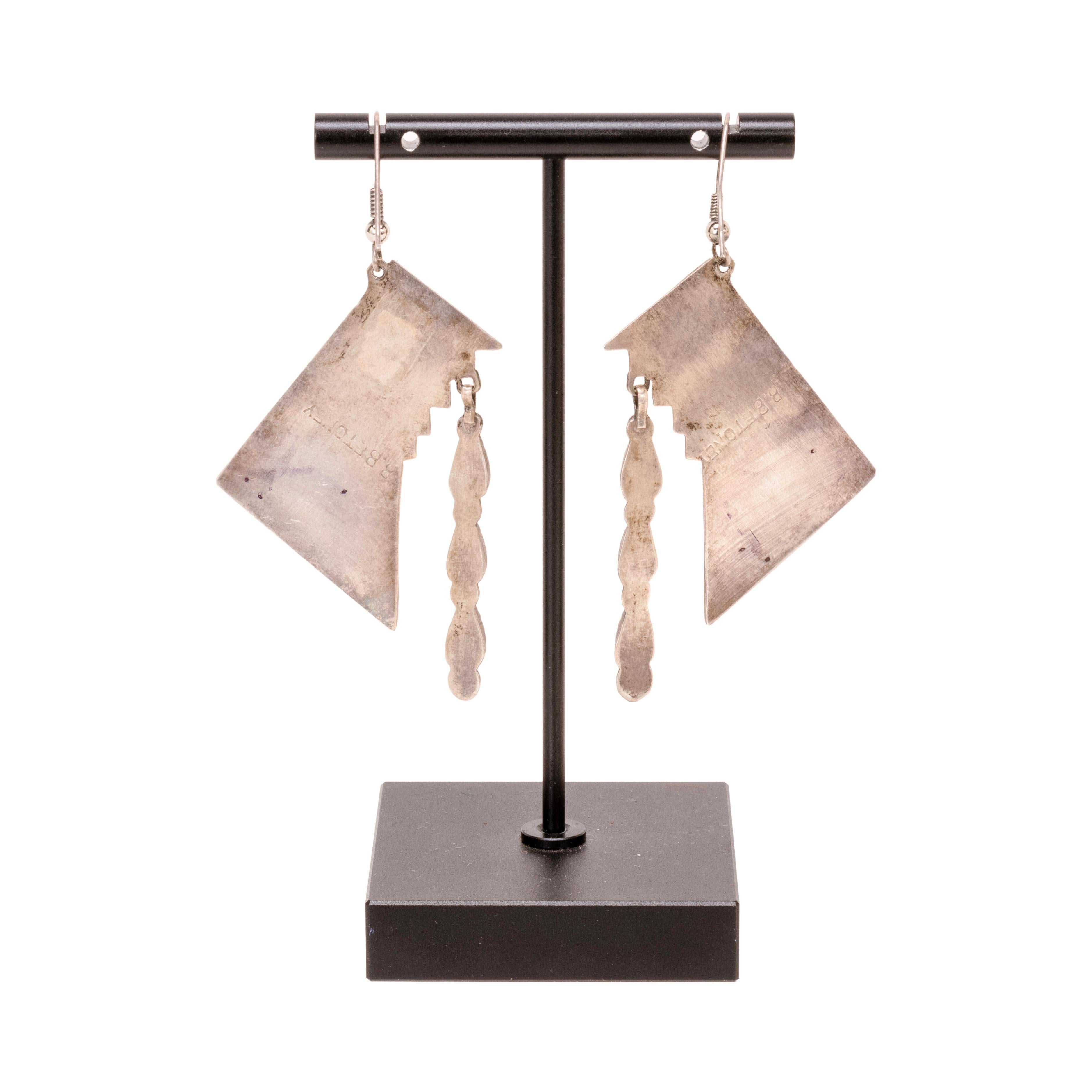 Zuni Ohrringe, hergestellt und markiert von V. Betone. Geometrischer Sockel aus Sterling mit geätzten Motiven. Einzelne tropfenförmige Korallenperle in der unteren Ecke vor schraffiertem Sterlinghintergrund. In der mittleren oberen Ecke baumelt ein